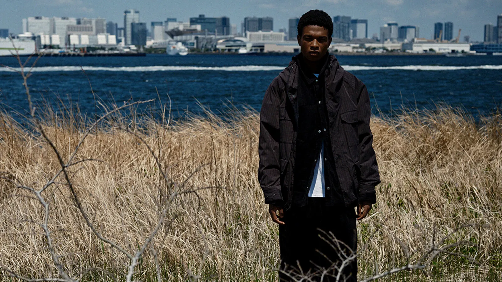 THE NORTH FACE PURPLE LABEL 마운틴 파카 착장 전신 컷 — 갈대밭과 바다, 도쿄 스카이라인을 배경으로 선 실루엣
